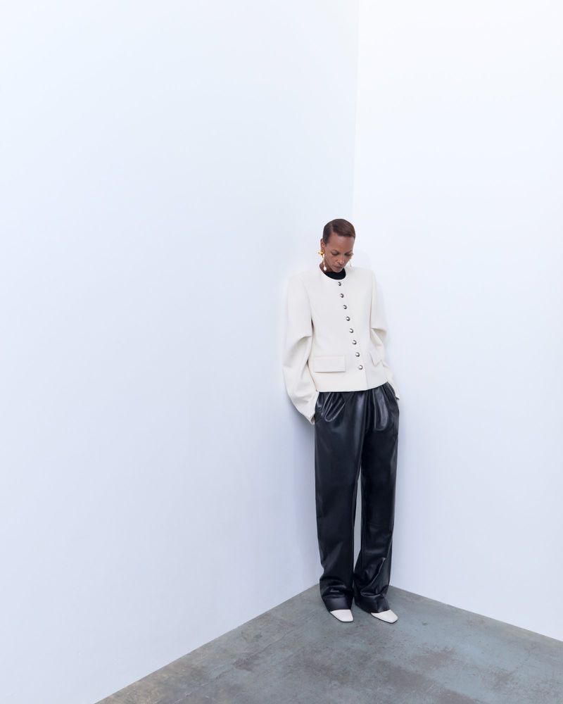 A person with short hair wearing a cream jacket and black pants poses against a stark white wall, looking downward in a minimalist fashion shot.