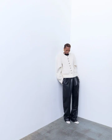 A person with short hair wearing a cream jacket and black pants poses against a stark white wall, looking downward in a minimalist fashion shot.