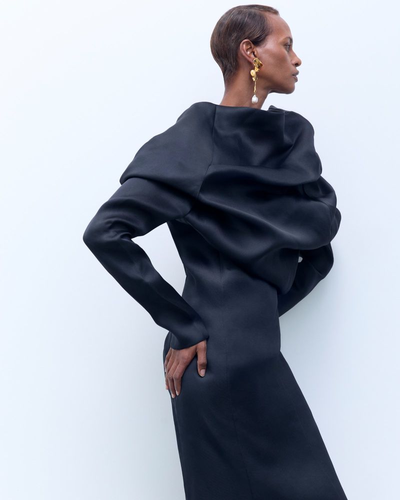Elegant woman poses in long-sleeved black dress with architectural shoulder detail against a bright neutral backdrop in studio setting.