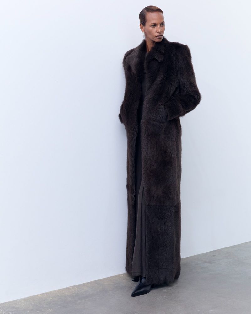 Model wearing a long dark brown fur coat standing against a plain white studio background.