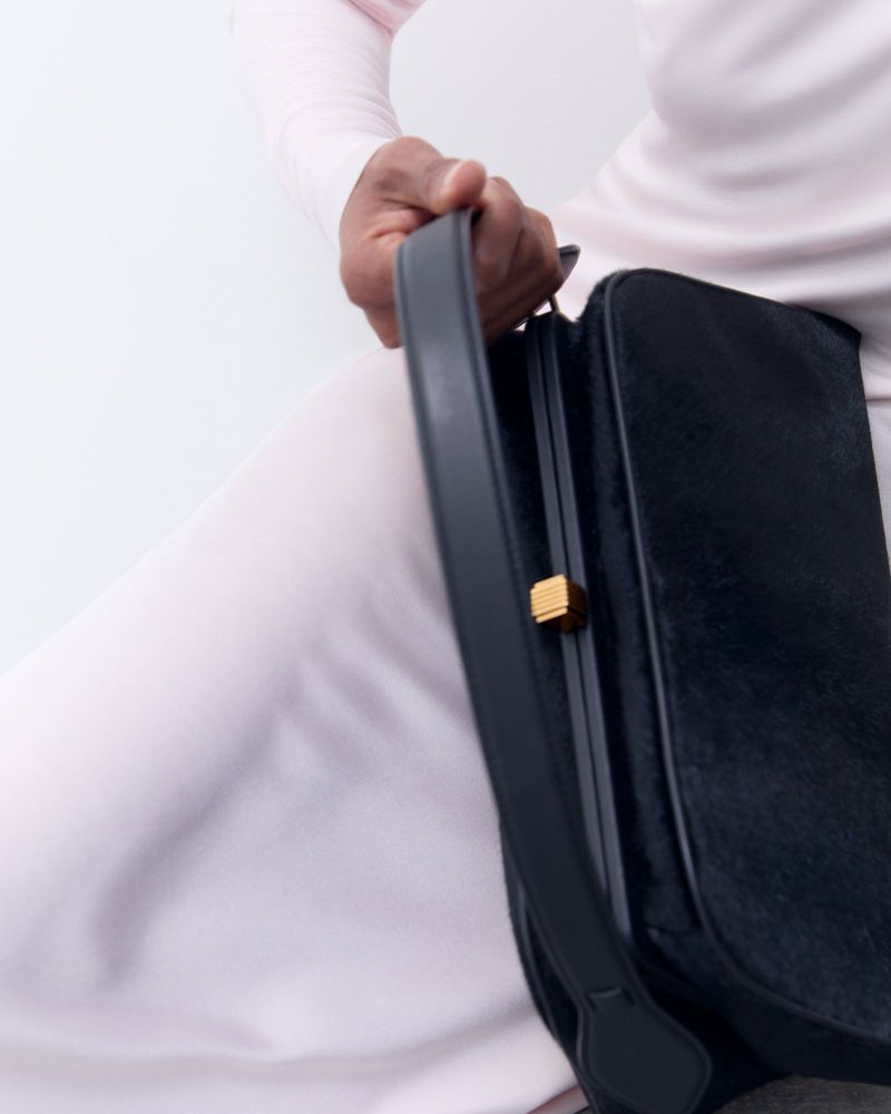 Model wearing a light pink long-sleeve top holding a black handbag with a gold closure against a white background.