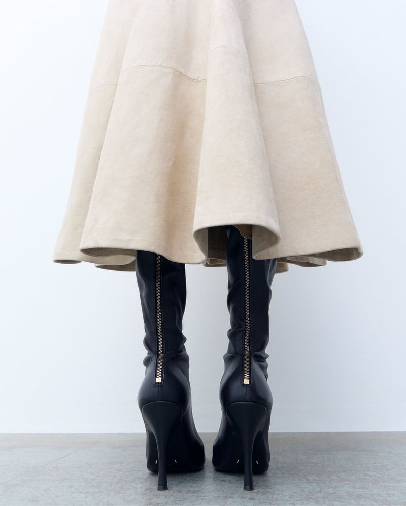 Woman wearing a flared tan skirt and black stiletto heel boots with zippers, standing against a plain white wall.