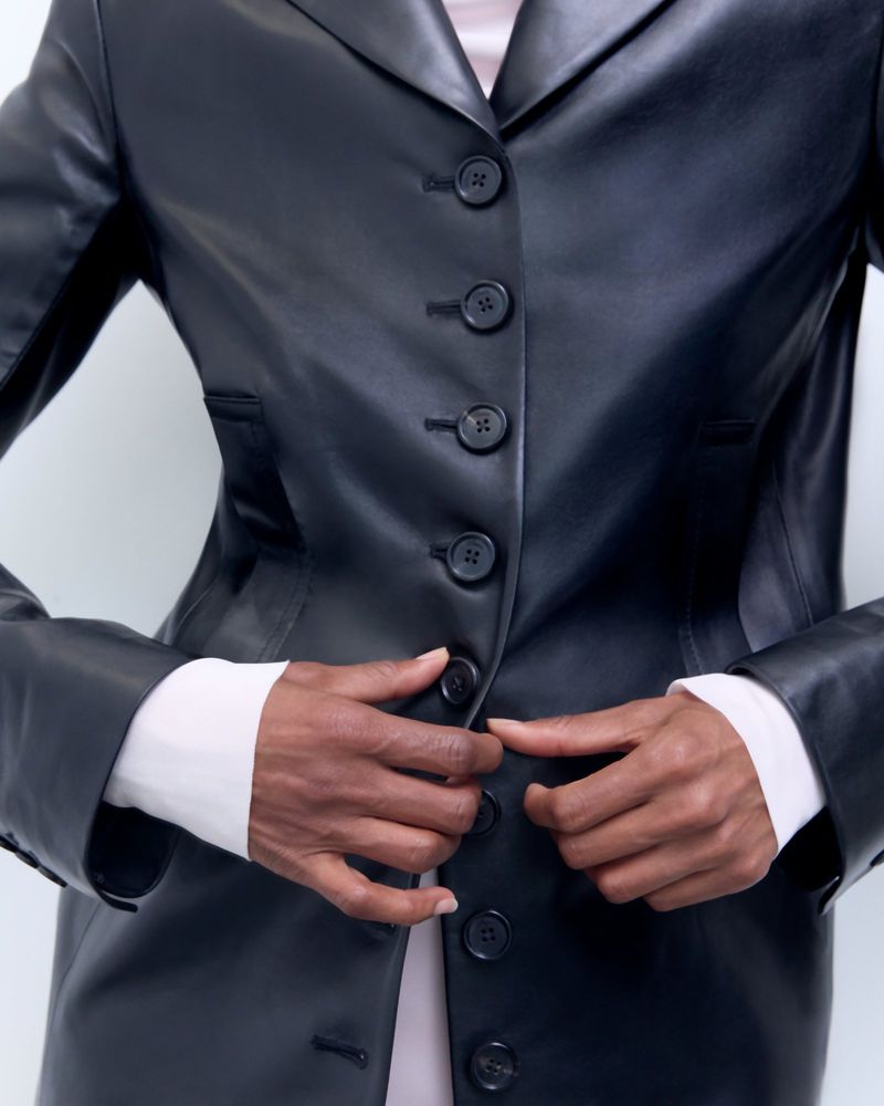Dark-skinned adult fastens buttons on a sleek, dark leather jacket with a light pink top underneath. Studio shot, upper body focus.