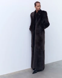 Model wearing a long dark brown fur coat standing against a plain white studio background.