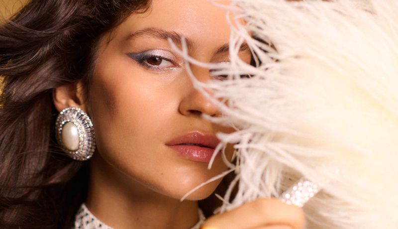 Close-up shot of a glamorous adult female model with dramatic eye makeup, wearing large earrings, and holding a white feather fan.