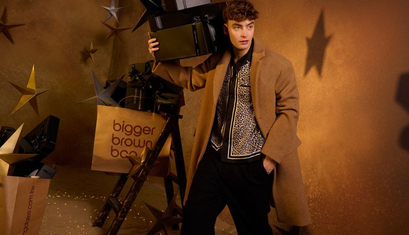A young adult male stands with shopping bags and gift boxes in a retail setting, adorned with star decorations and warm lighting.