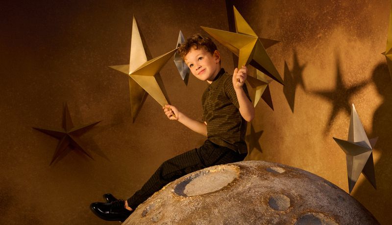 Boy sits on a moon prop holding star props in a studio against a backdrop of stars, smiling into the camera with a warm, inviting light.