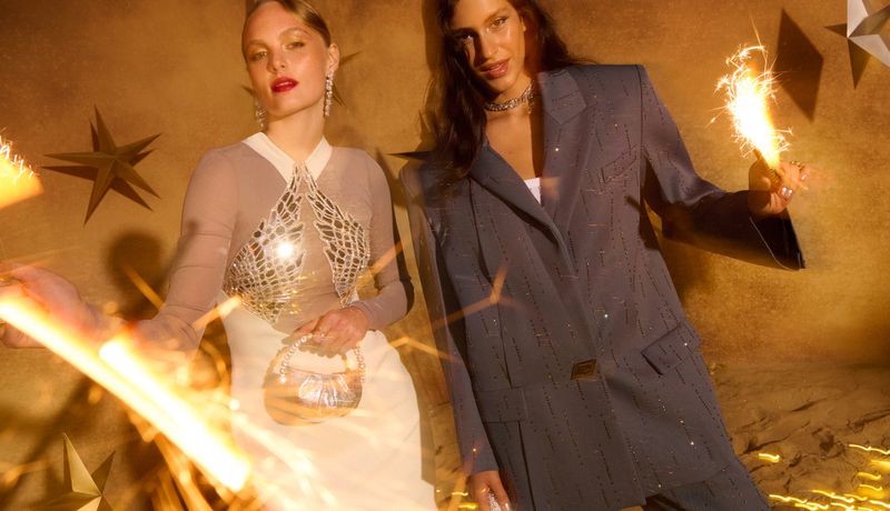 Two women pose with sparklers in a studio with star decorations. One wears a pinstripe suit and the other a silver embellished top and white skirt.