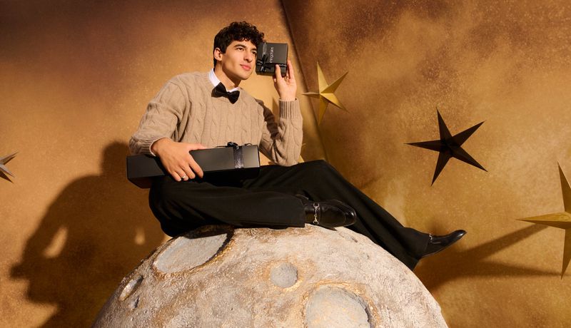 Adult male sits on a moon prop, holding a gift box and camera against a celestial backdrop with stars, wearing a tan sweater and bow tie.