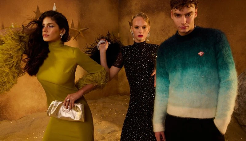 Three fashion models pose in a stylized indoor environment with textured sand and star decorations, showcasing formal evening wear.