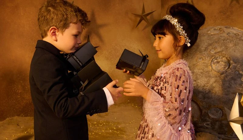 A young boy in a suit presents a gift to a girl in a sparkly dress in a staged environment with a moon and stars backdrop.