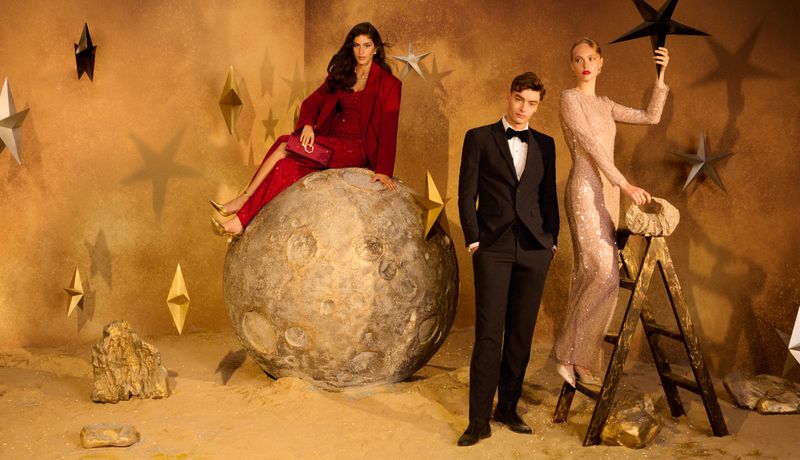 Two women and one man in formal wear pose with a moon prop and star decorations in a warm-toned studio setting.