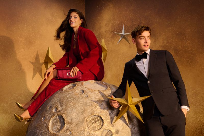Glamorous couple poses with stars and a moon prop. The woman wears a red suit and gold heels, the man a tuxedo with a bowtie.