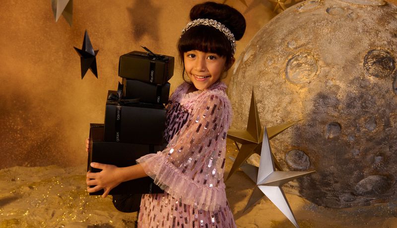 Young girl in pink dress carries stack of black gift boxes against moon and star backdrop in studio with warm, golden lighting.