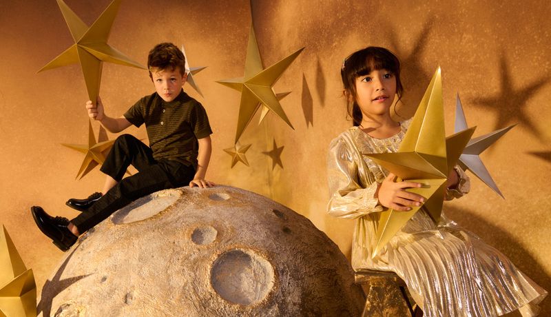 Two children pose on a moon prop holding gold star props against a gold backdrop for a playful, creative, celestial themed photograph.