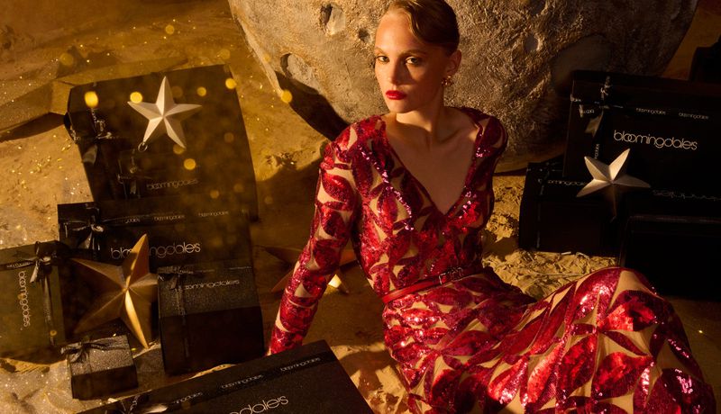Model in red sequin dress seated among branded shopping boxes and star props in a festive holiday campaign image.