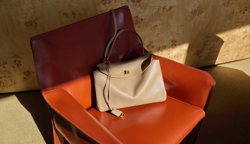 A tan leather handbag with a top handle and a shoulder strap rests on a red leather chair against a patterned wooden wall backdrop.