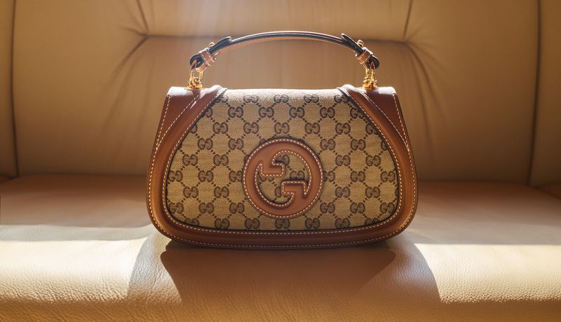 Gucci handbag on tan leather couch, displaying the brand's iconic monogram pattern with a circular logo and a structured handle.