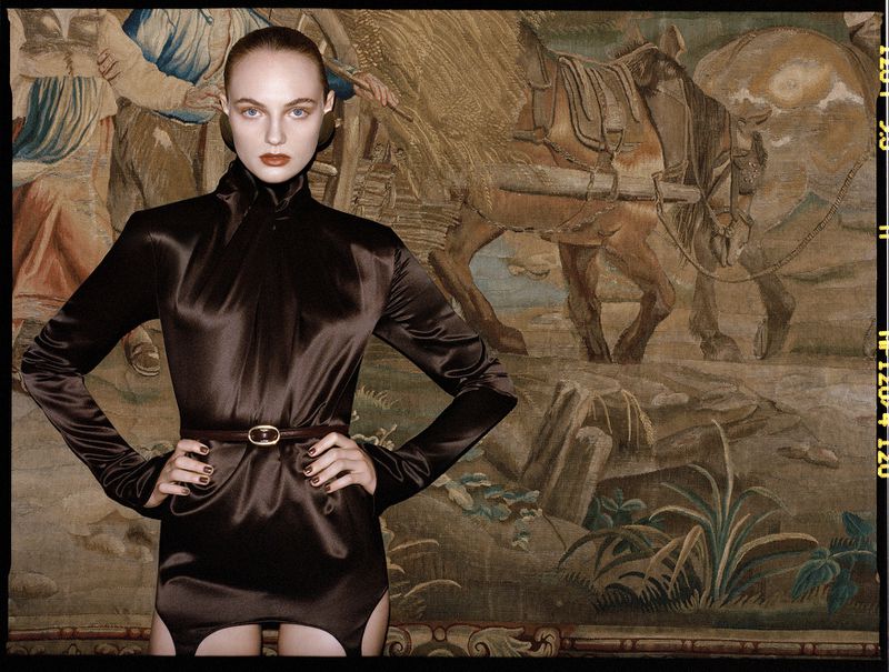 Fashion editorial shot of a fair-skinned woman in a brown bodysuit with a belt, posing in front of a tapestry featuring horses.