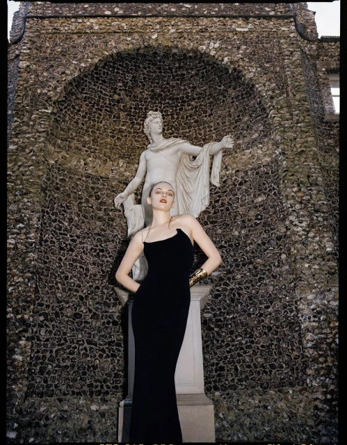 Elegant woman in a black gown poses in front of a stone statue and textured stone wall with an arched alcove, outdoors.