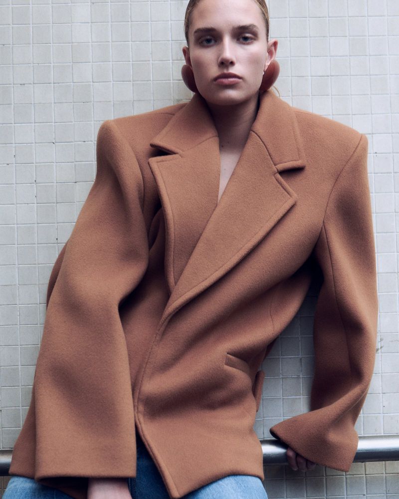 Fashion shot of a young adult female wearing a brown wool blazer and jeans in an indoor setting with a tiled wall backdrop.