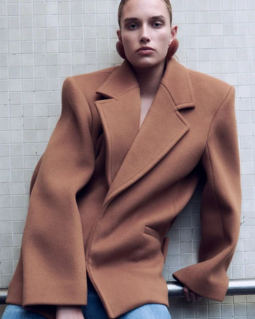 Fashion shot of a young adult female wearing a brown wool blazer and jeans in an indoor setting with a tiled wall backdrop.