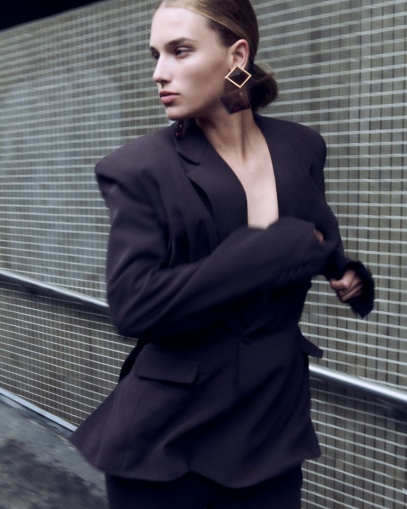 Fashion shot of a woman in a dark blazer and square gold earrings against a grey tiled wall, showcasing modern elegance.
