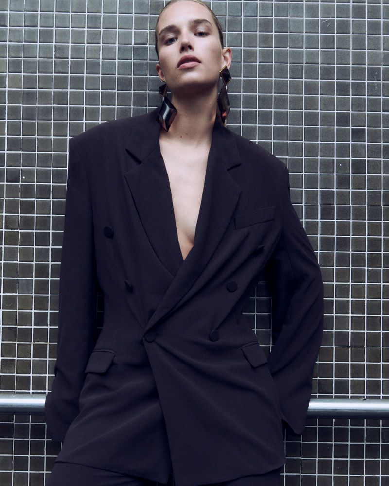 A woman in a black blazer and earrings poses against a grid wall, creating a sophisticated fashion image with a modern edge.