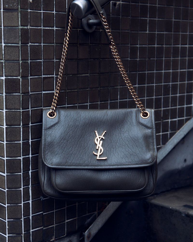 Elegant YSL shoulder bag with a gold chain, hanging against a backdrop of dark brown tilework, creating a stylish and luxurious aesthetic.