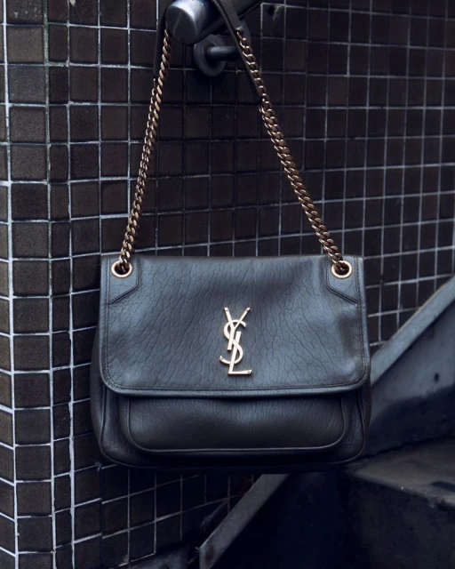 Elegant YSL shoulder bag with a gold chain, hanging against a backdrop of dark brown tilework, creating a stylish and luxurious aesthetic.