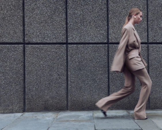 Adult woman in a taupe pantsuit strides confidently on a sidewalk alongside a textured concrete wall, showcasing a blend of fashion.