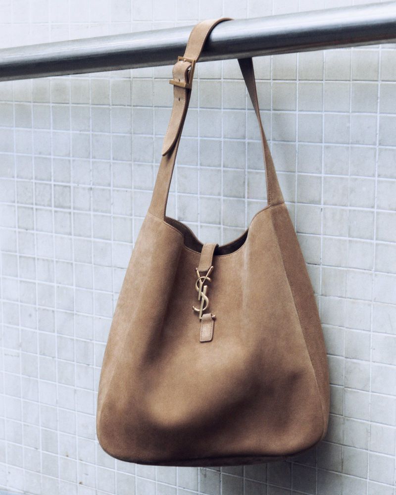 Tan suede Saint Laurent hobo bag hanging from a metal bar against a textured white tile background. Fashion accessory shot.