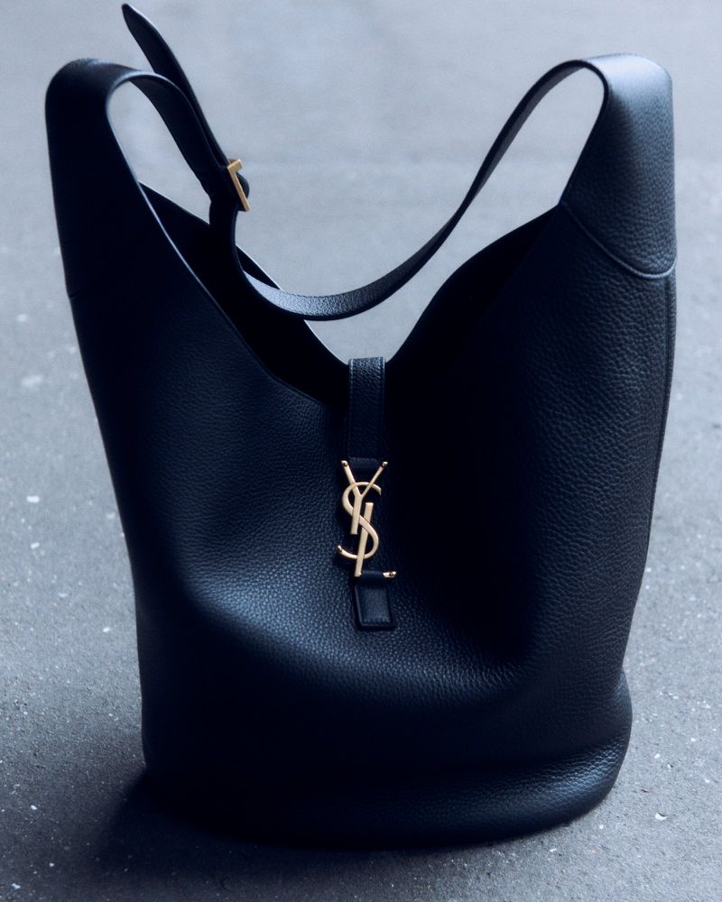 Black leather bucket bag with a gold YSL logo, featuring a single strap and resting on a grey concrete surface. Fashion.