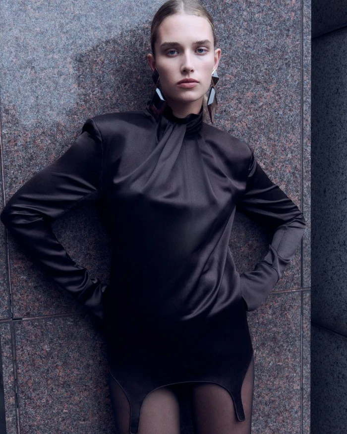 Model wearing a black satin high neck dress posing against a dark mottled stone wall for Net-A-Porter editorial.