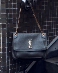 Dark grey leather shoulder bag with gold chain strap and interlocking YSL logo hanging against dark tiled wall