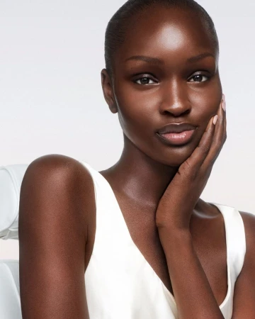 Close-up portrait of a dark-skinned woman with hand on face, wearing a white top, against a soft white background in a beauty shot.