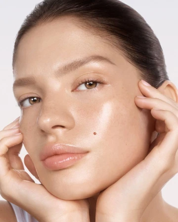Close-up portrait of a young adult woman with light skin and brown hair, posing with her hands near her face against a neutral background.