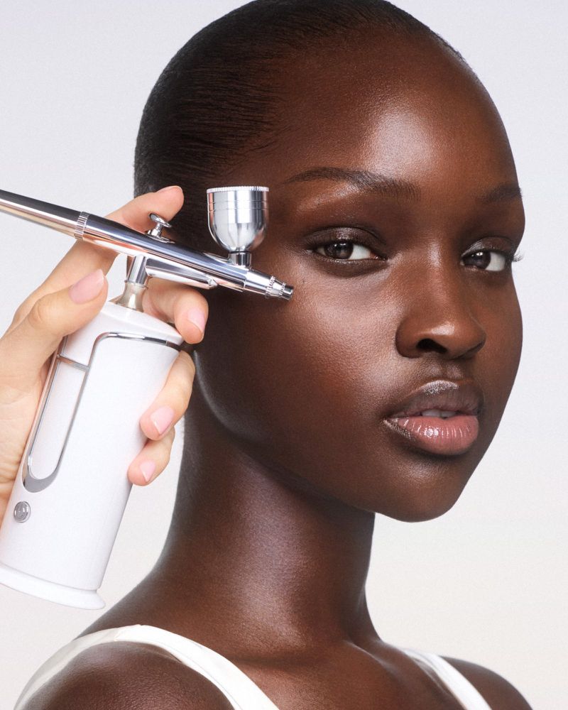 A dark-skinned woman receiving an airbrush facial treatment indoors, with a close-up showing the device and her face.