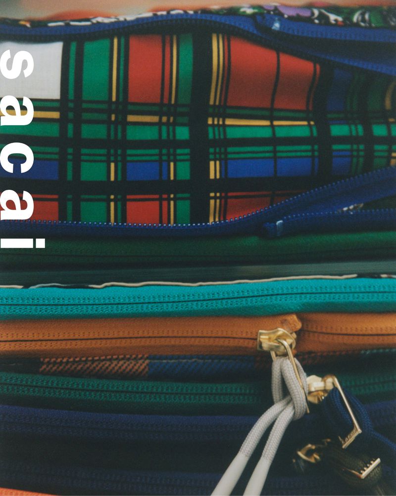 Close-up of a stack of multicolored zippered pouches with plaid pattern and a rope detail, with the text 'sacai' on the side.