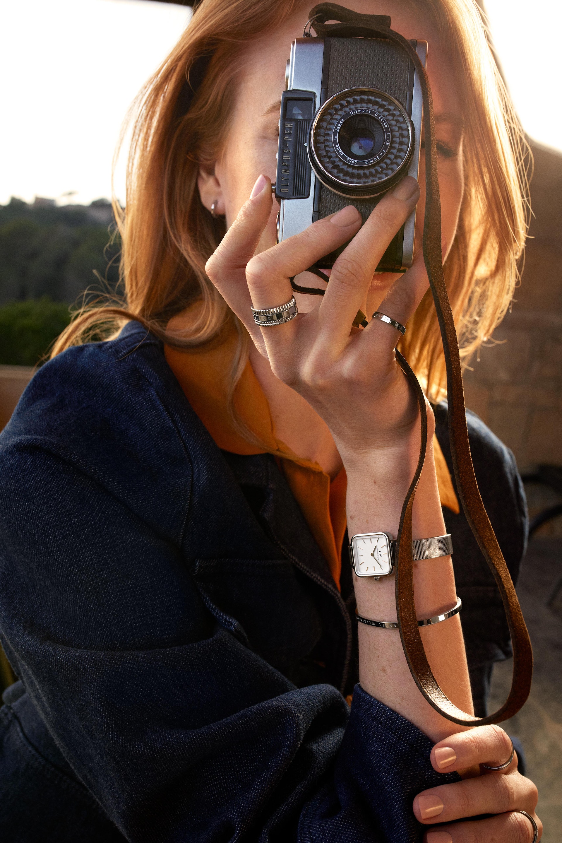Young woman with red hair takes a photo with a vintage camera outside, wearing a denim jacket and silver jewelry in golden sunlight.