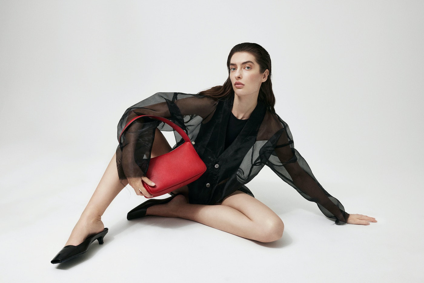 Studio shot of a woman with a red purse. The model is sitting on the floor wearing a sheer black jacket and black shoes.