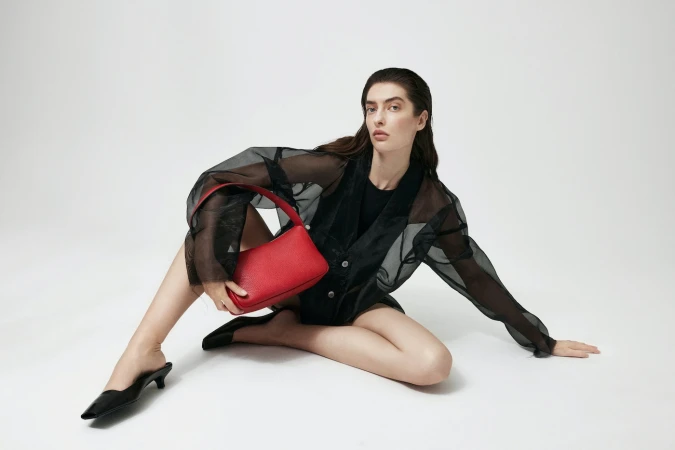 Studio shot of a woman with a red purse. The model is sitting on the floor wearing a sheer black jacket and black shoes.