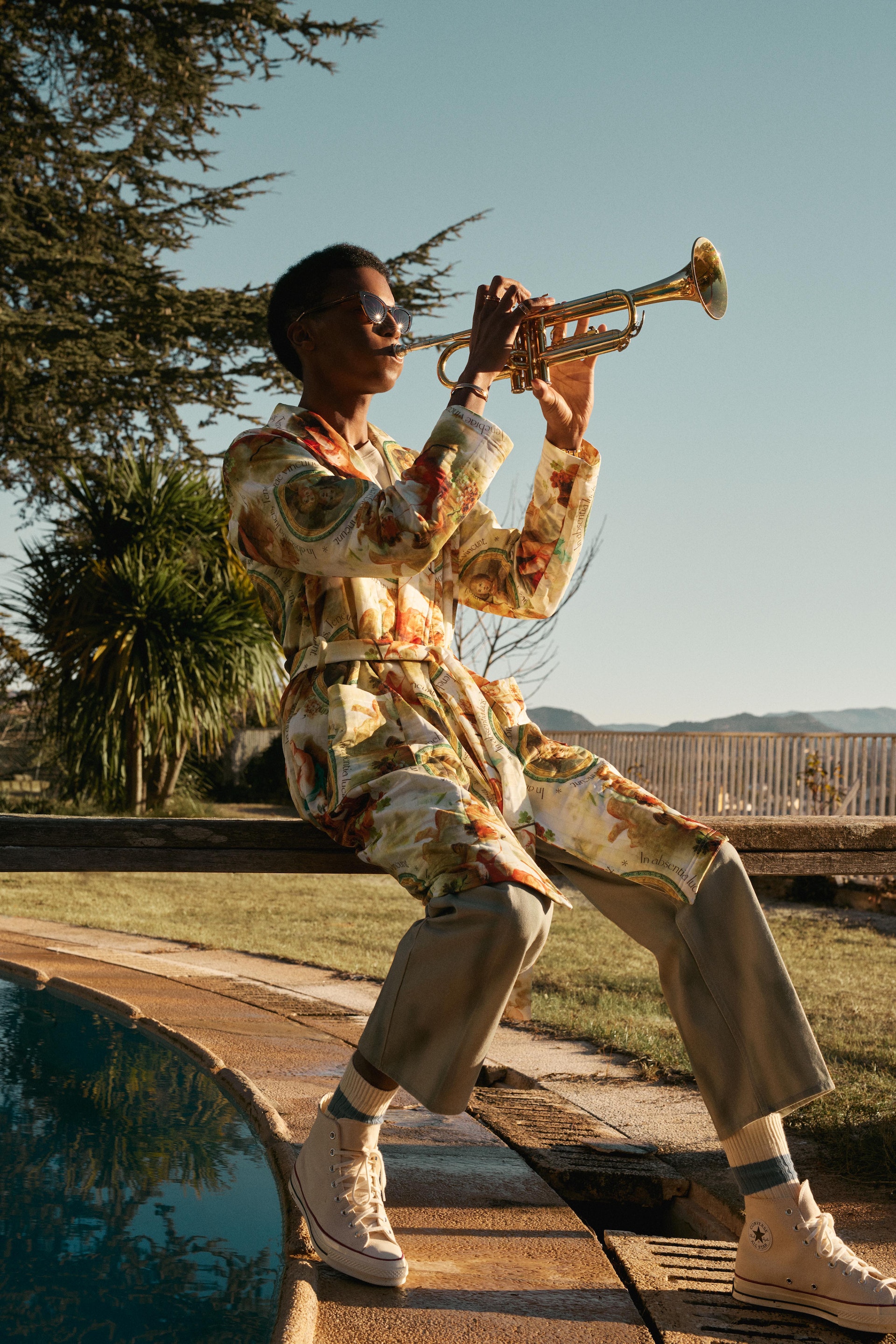 A person plays a trumpet outdoors. The individual wears a floral robe, cropped pants, and sneakers, sitting on a ledge near a pool with trees nearby.