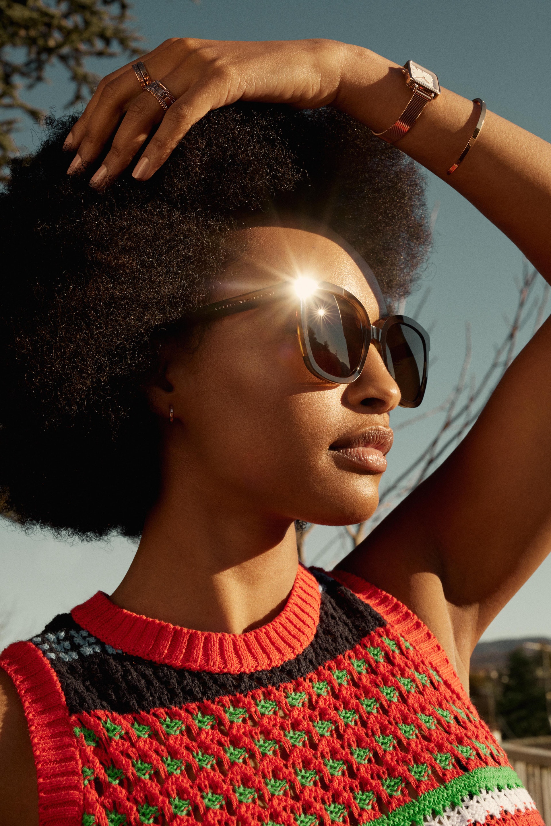 Outdoor portrait of an adult woman with afro hairstyle wearing sunglasses and a colorful knit top on a sunny day.