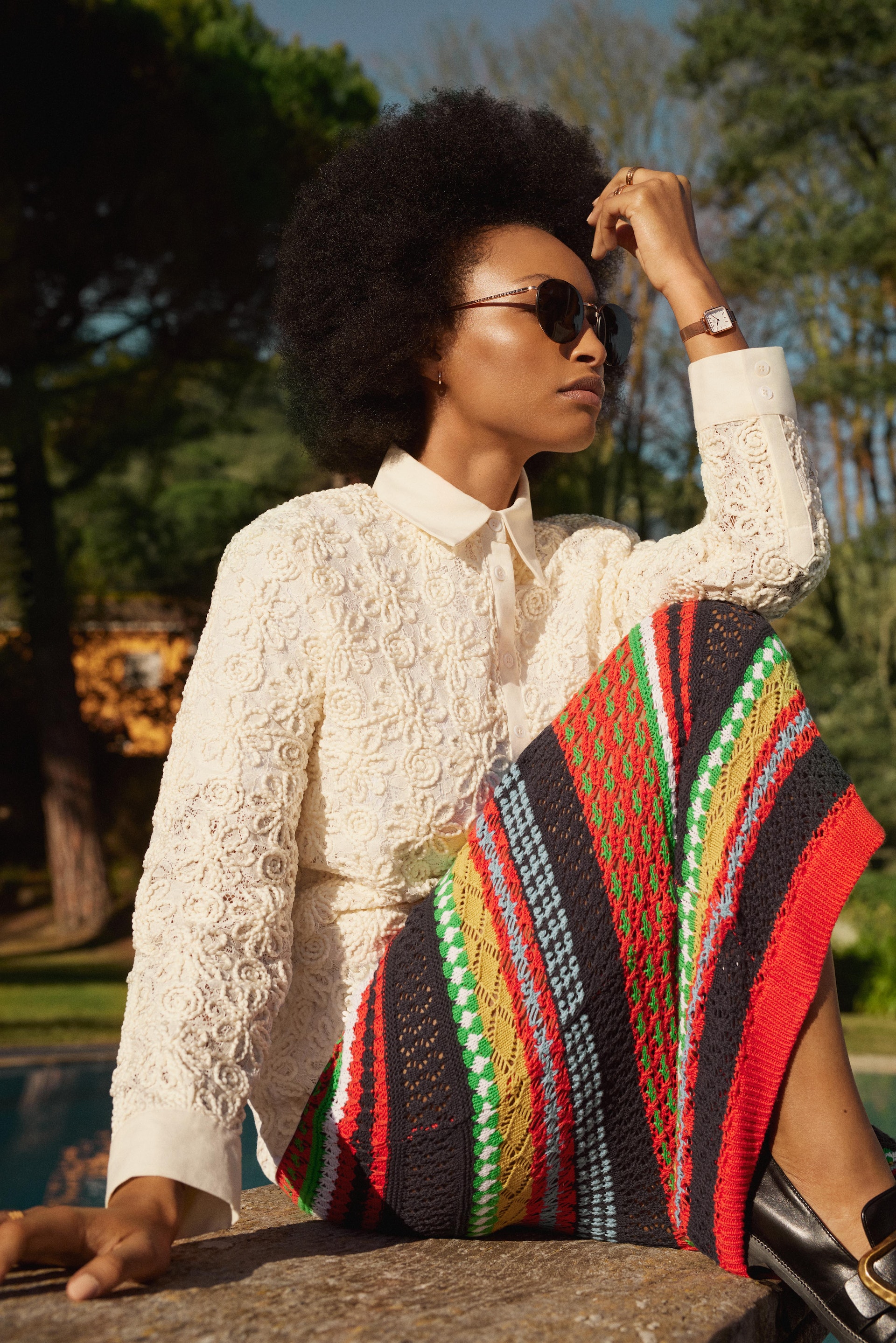 A stylish woman with an afro sits outdoors, wearing sunglasses, a white floral lace shirt, and a colorful patterned skirt in daylight.