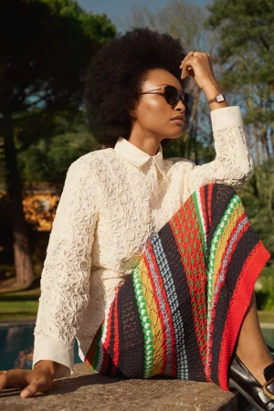 A stylish woman with an afro sits outdoors, wearing sunglasses, a white floral lace shirt, and a colorful patterned skirt in daylight.