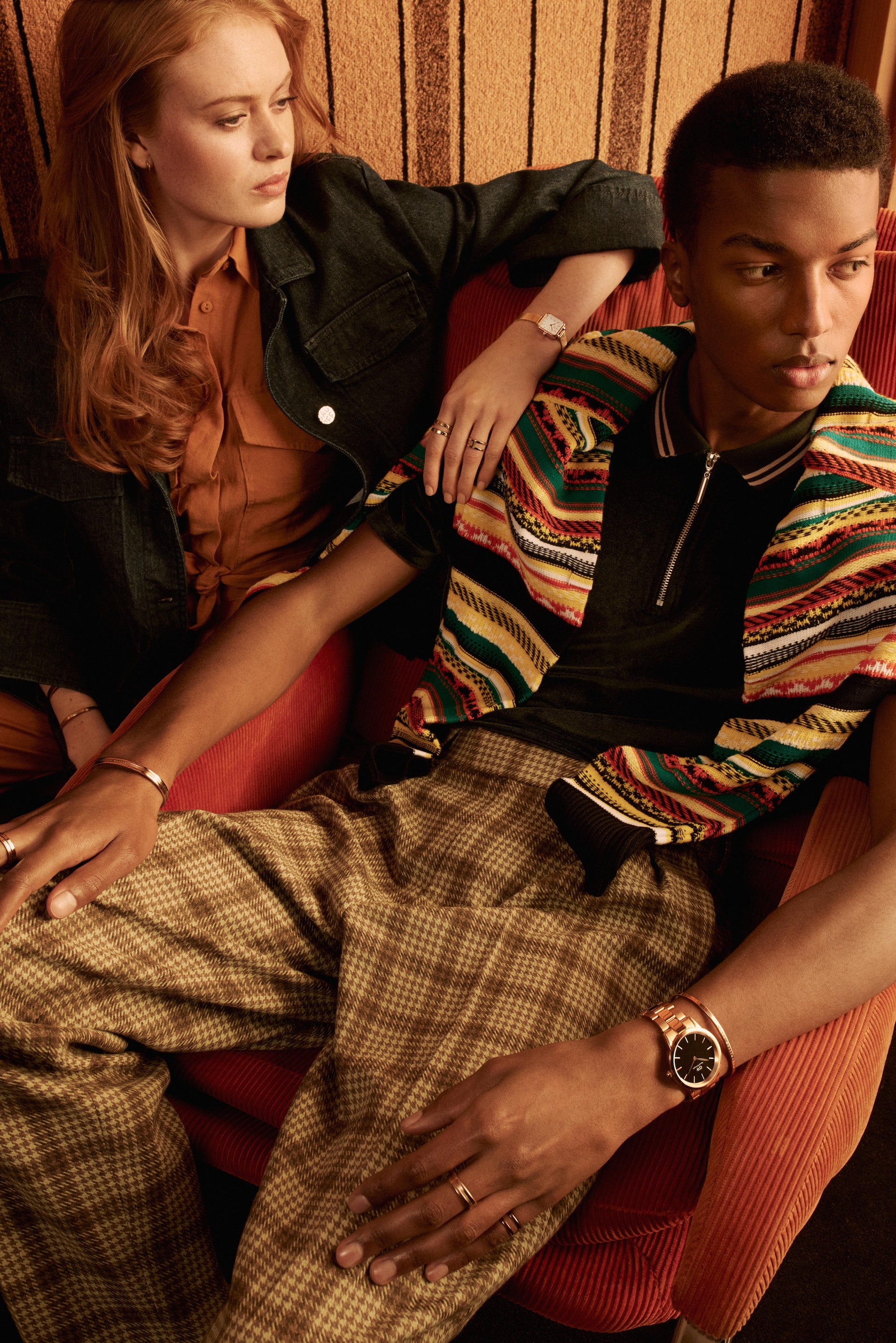 A stylish couple is photographed indoors, showcasing complementary fashion styles and jewelry against a striped backdrop, with warm, natural lighting.