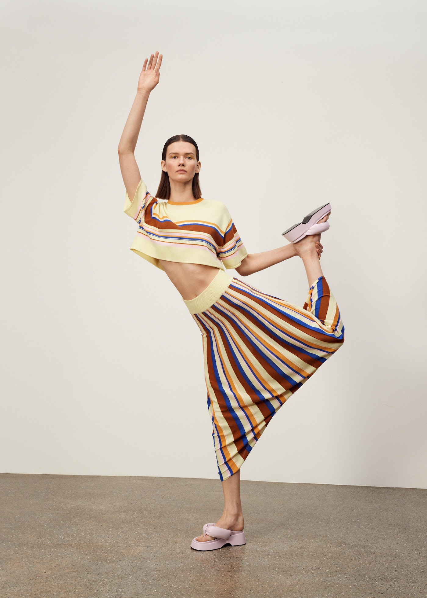 A fair-skinned adult woman balances in a yoga pose indoors, modeling a striped crop top, striped skirt, and platform sandals in a studio shot.