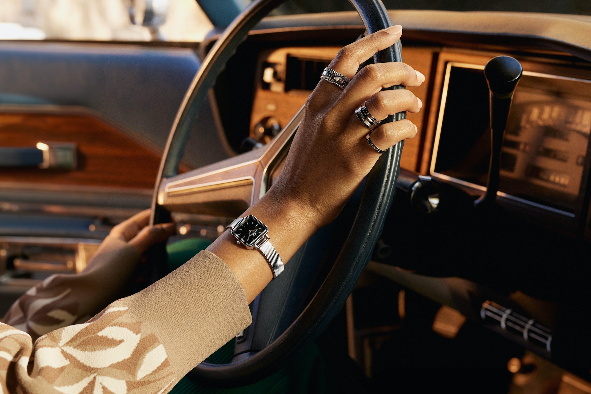 Close-up shot inside a vintage car: a hand on the steering wheel wearing a wristwatch and rings, in golden light.