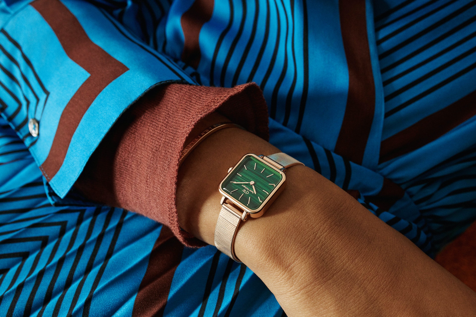 Close-up shot of a wrist wearing a gold-toned, square-faced watch with a green malachite dial, accented by a striped blue and brown shirt.